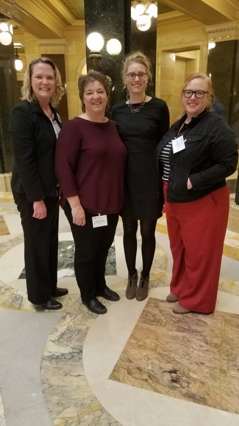 Four female staff members dressed in business attire smiling inside the Madison Capitol building
