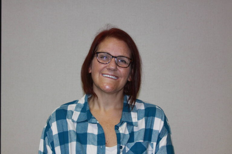 Woman smiling with dark red hair, glasses, and blue and white plaid shirt