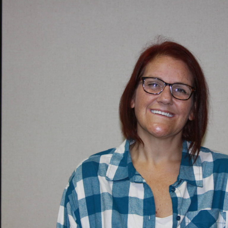 IMG_9141 Woman smiling with dark red hair, glasses, and blue and white plaid shirt