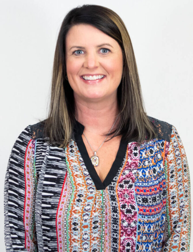 Headshot of woman with straight shoulder length brown hair and highlights, smiling at camera with patterned top