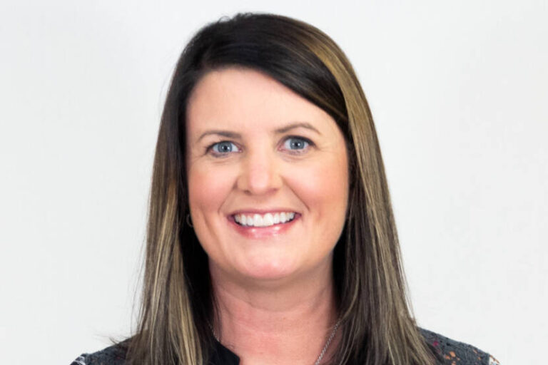 Headshot of woman with straight shoulder length brown hair and highlights, smiling at camera with patterned top