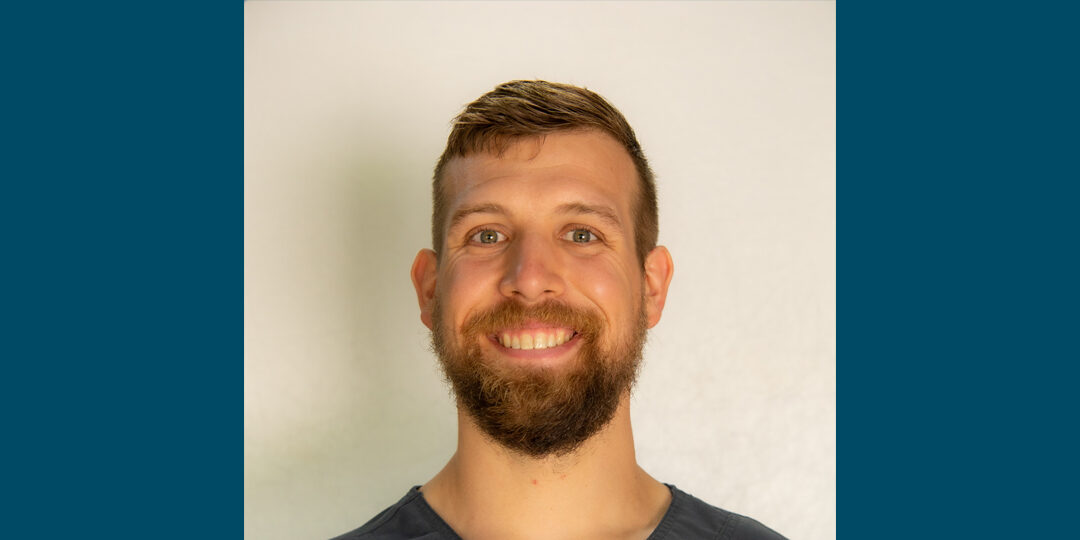 Headshot of man with dark gray scrub top and beard, smiling