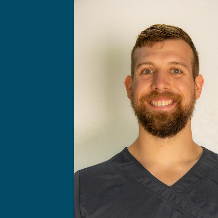 Benjamin-Tenpas-1080×720 Headshot of man with dark gray scrub top and beard, smiling