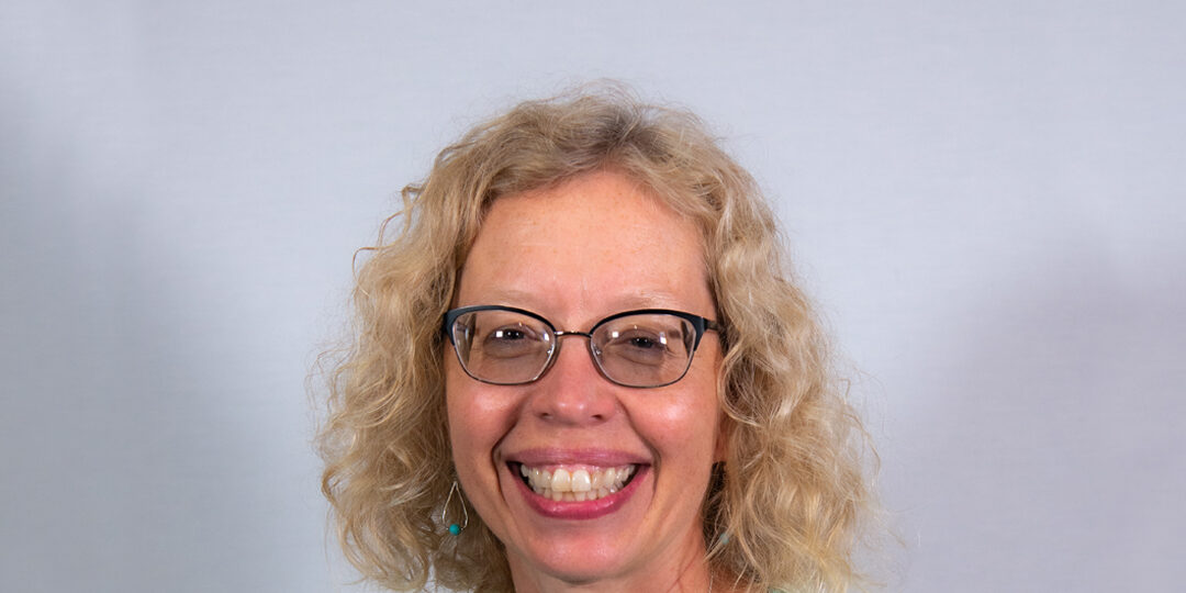 Rebecca-Crumb-Johnson-1080×720 Headshot of woman with blond curly haired woman with light blue tank top