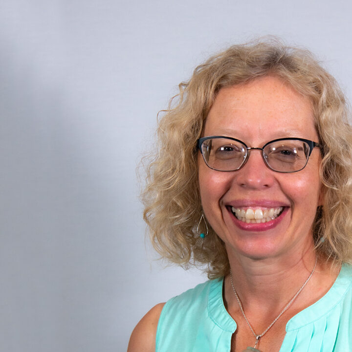 Rebecca-Crumb-Johnson-1080×720 Headshot of woman with blond curly haired woman with light blue tank top