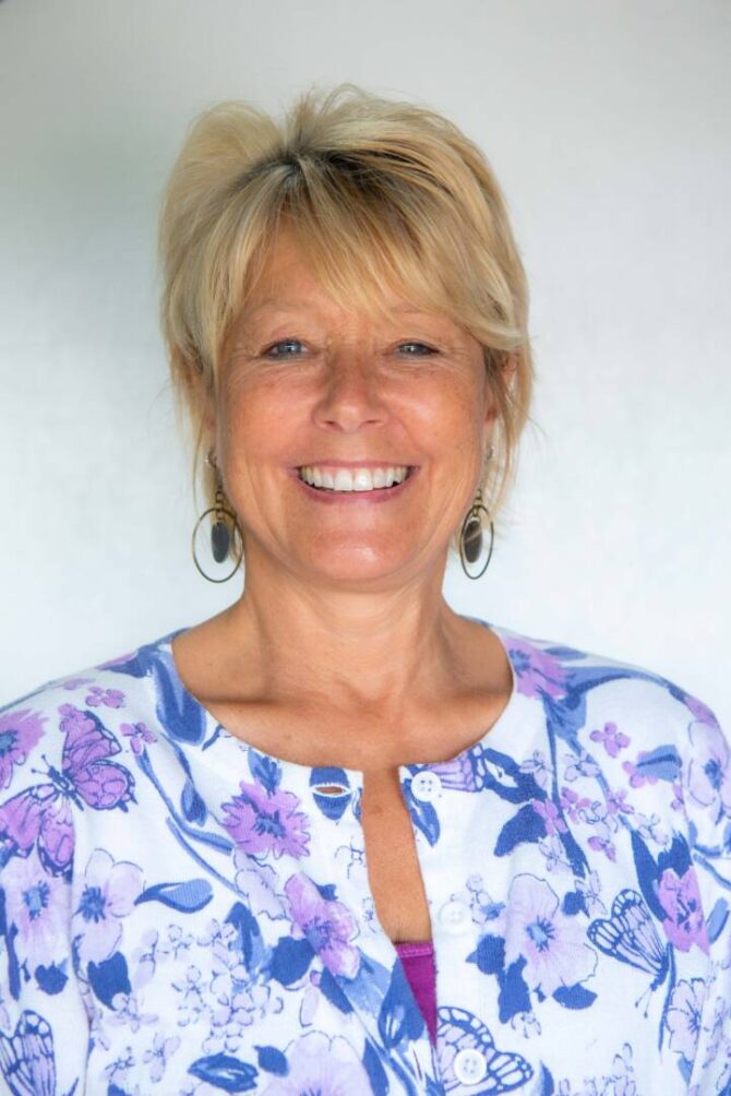L.WakefieldWeb 08-2021 Headshot of woman with blonde ponytail smiling at camera with white and purple bright floral top