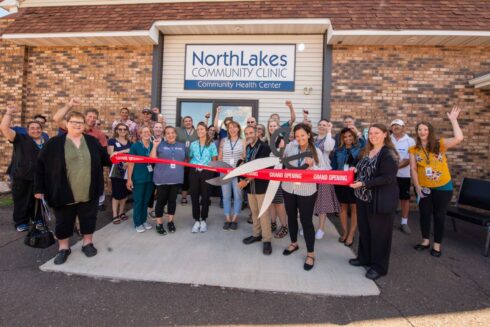 Large group of men and women standing outside of brick clinic building. One woman is holding a large pair of scissors to cut a red ribbon.