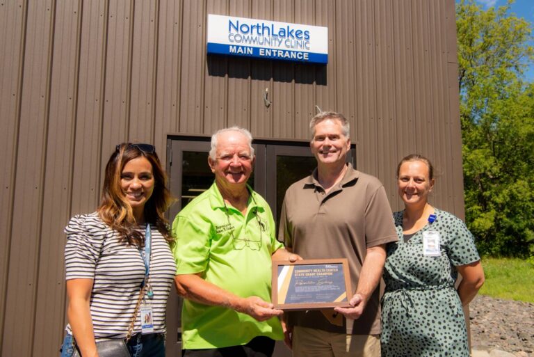 4 people standing holding "Community Health Center State Grant Champion" sign in front of Hayward Clinic.