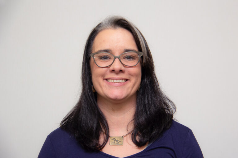 Headshot of woman smiling with shoulder length black hair and blond highlight, wearing glasses