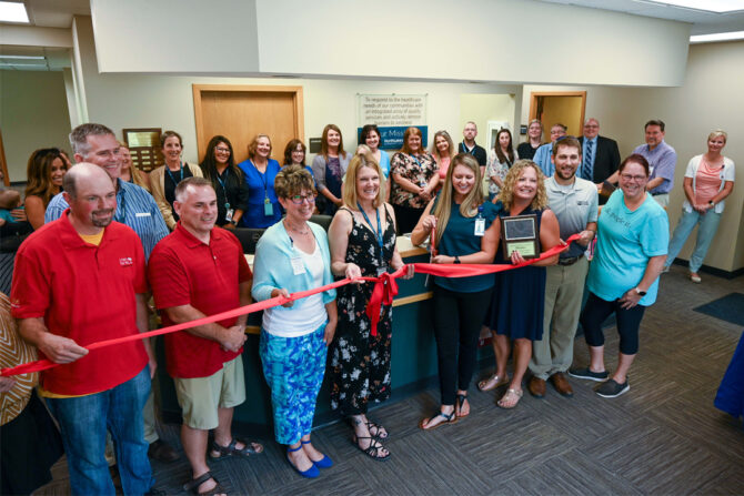 Cumberland-Ribbon-Cutting-Ceremony-Featured-Image Large group of men and women standing and holding a red ribbon in a clinic lobby