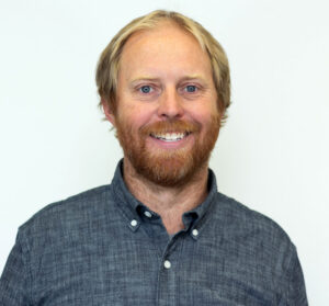 Headshot of man with blond hair and beard wearing gray button up long sleeve