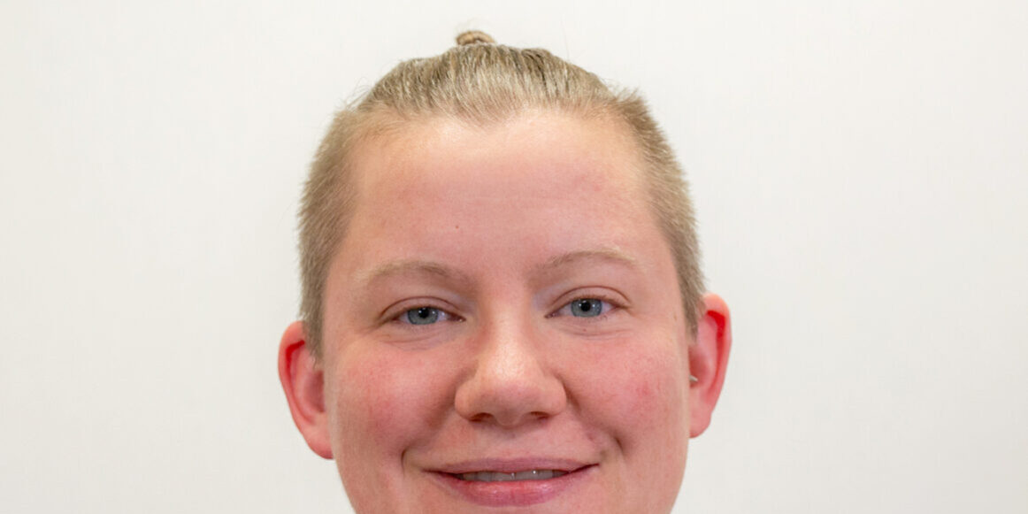 Headshot of person with navy blue waffle shirt, light blue eyes, and blonde hair pulled back into small bun