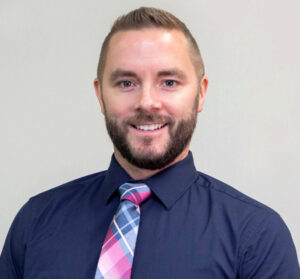 Headshot of man with short brown hair and facial hair wearing a dark blue dress shirt and plaid tie