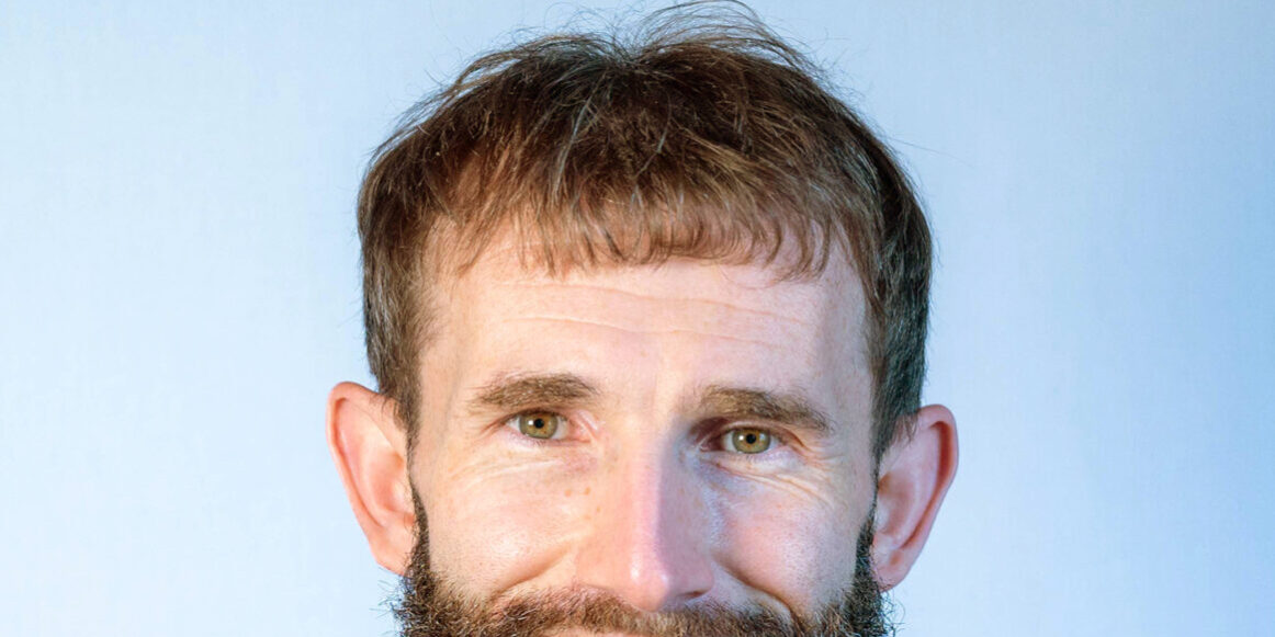 Man with brown hair and beard wearing green and black plaid shirt