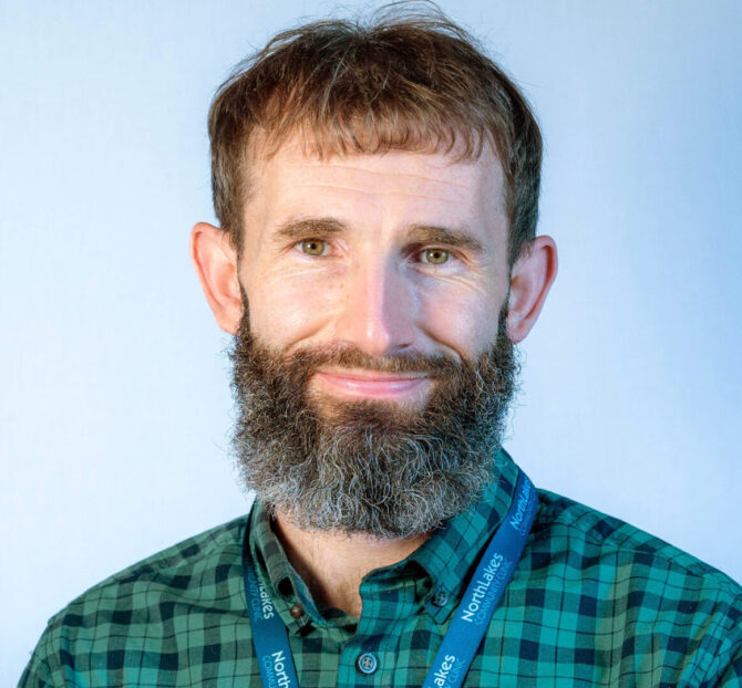 Man with brown hair and beard wearing green and black plaid shirt