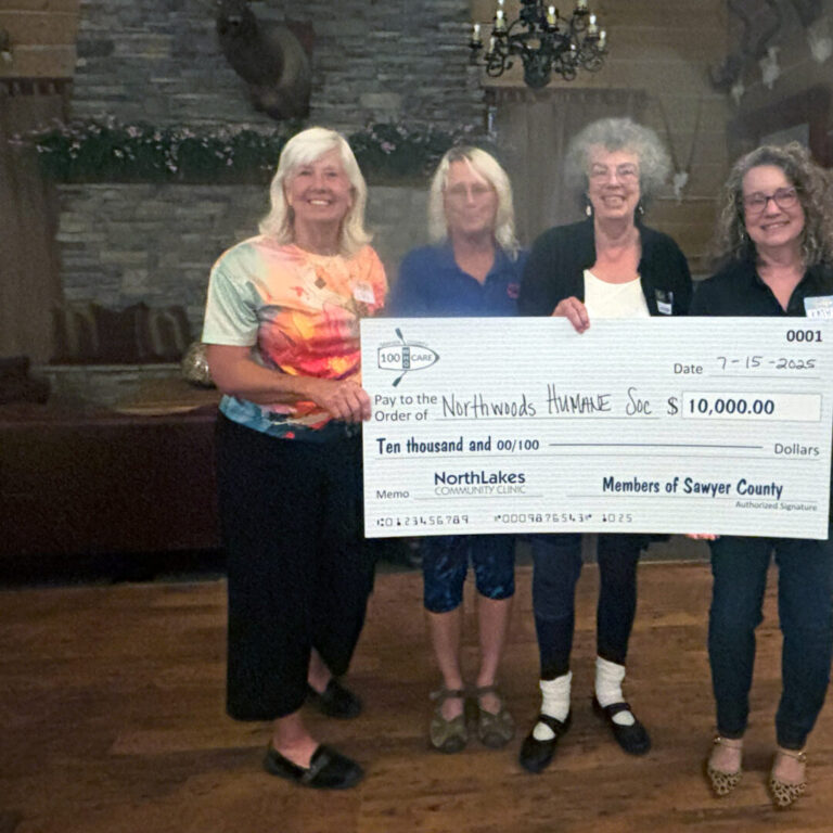 NorthLakes-Sawyer-100-who-care Four people holding a big check in front of a fire place