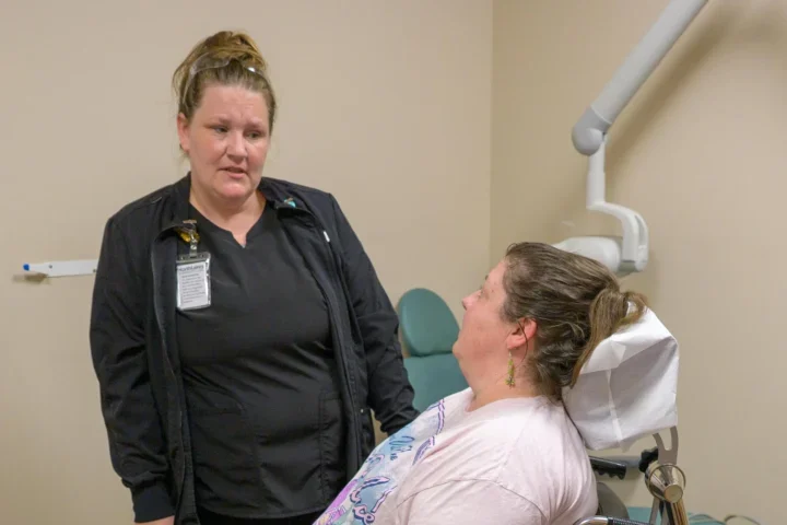 Two people speaking, one standing, and one using a new specialized wheelchair recliner at a NorthLakes dental clinic.